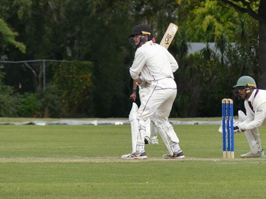 A cricket player in white gear prepares to bat, with a teammate positioned behind the stumps. The scene is set on a grassy field surrounded by trees.