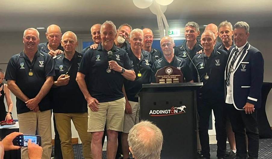 A group of older male cricket players celebrating at a podium with a trophy, wearing team jerseys and medals, during a tournament presentation.